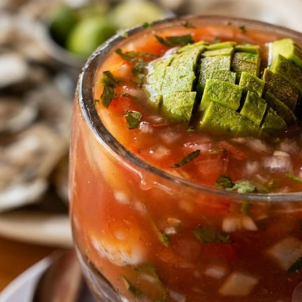 Close-up of a glass of red ceviche topped with diced avocado, with oysters in the background.
