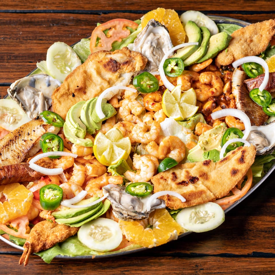 A large platter of seafood with shrimp, oysters, fried fish, avocado, and citrus on a wooden table.