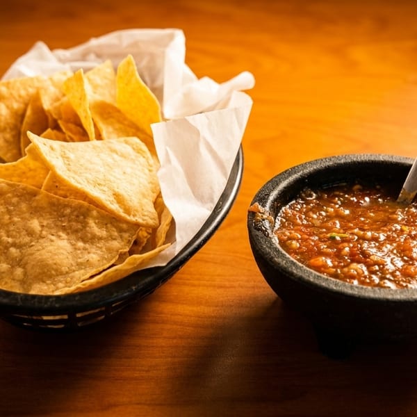 A basket of tortilla chips with a molcajete bowl of salsa and a spoon on a wooden table.