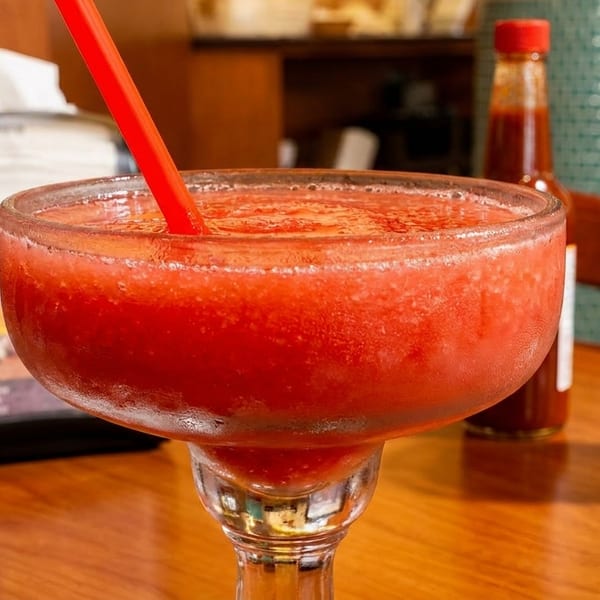 A vibrant red frozen margarita with a straw sits on a wooden table, with condiments in the background.