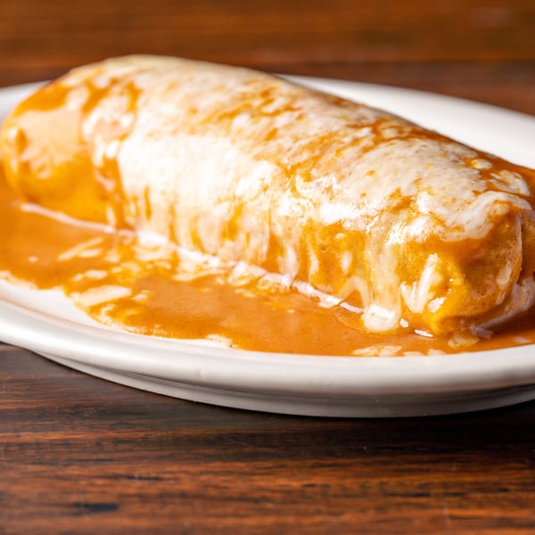 A large, cheesy burrito covered in red sauce on a white oval plate, on a wooden table.