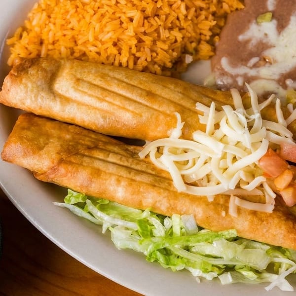 Two golden taquitos on lettuce with rice, refried beans, guacamole, and shredded cheese.