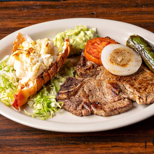 Grilled steak and lobster tail with guacamole, tomato, onion, and pepper on a white plate.